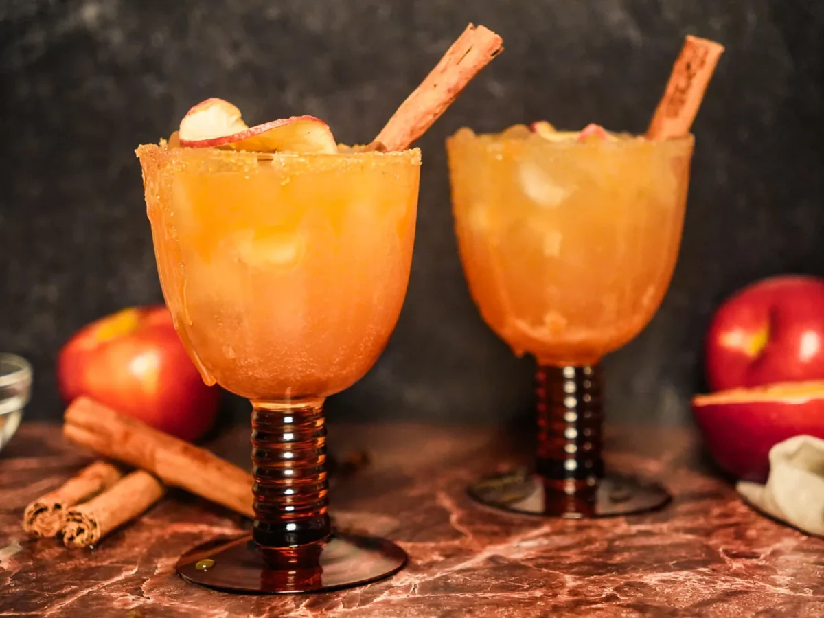 Two apple cider margaritas with cinnamon-sugar rims, dried apple slices, and cinnamon sticks, styled with fresh apples and cinnamon