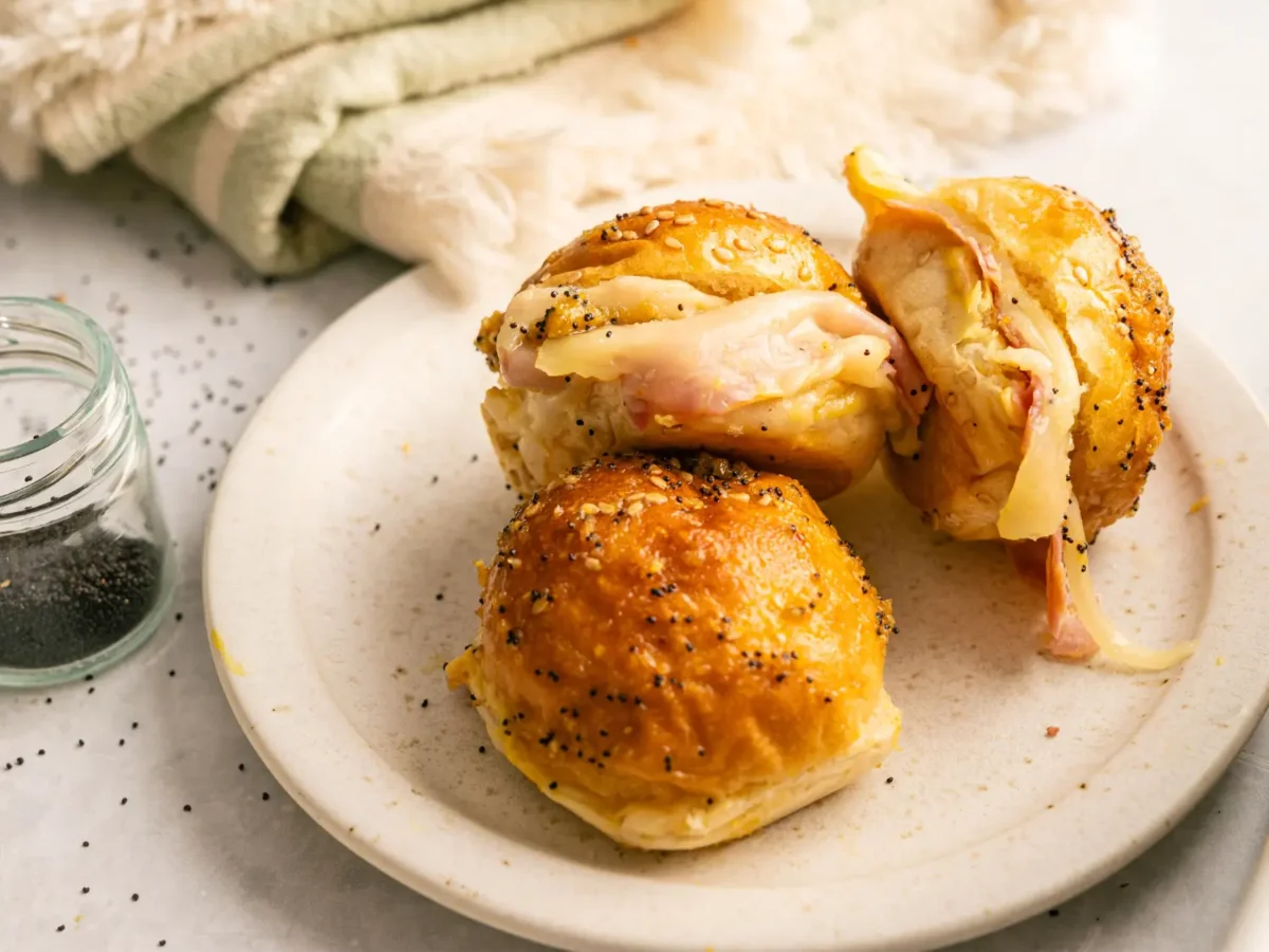 photo of three Hawaiian Bread Ham and Cheese Sliders on a cream plate next to a container of poppy seeds