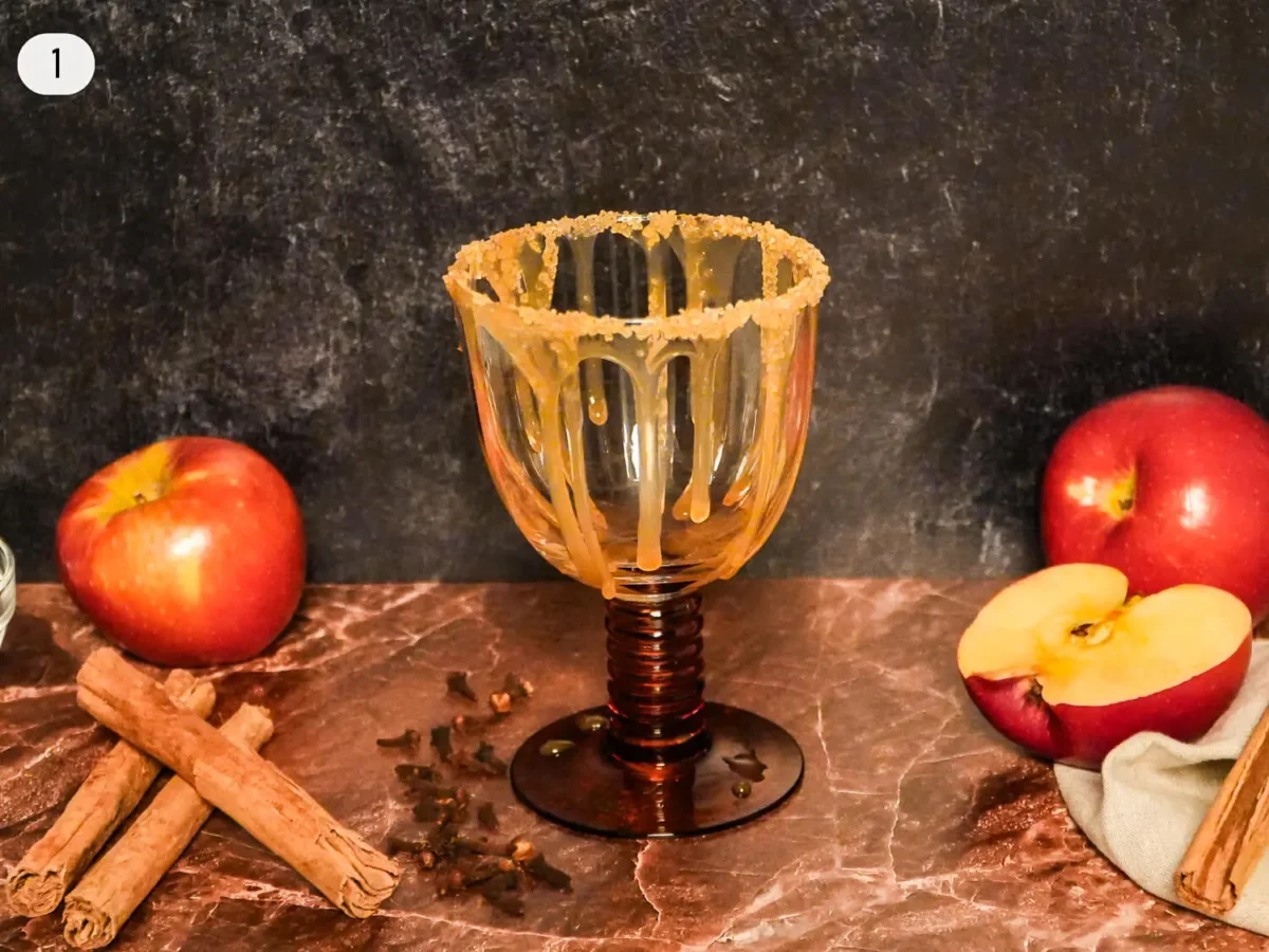 step 1 photo showing an empty amber cocktail glass prepared with a cinnamon-sugar rim and styled with fresh apples, cinnamon sticks, and whole spices