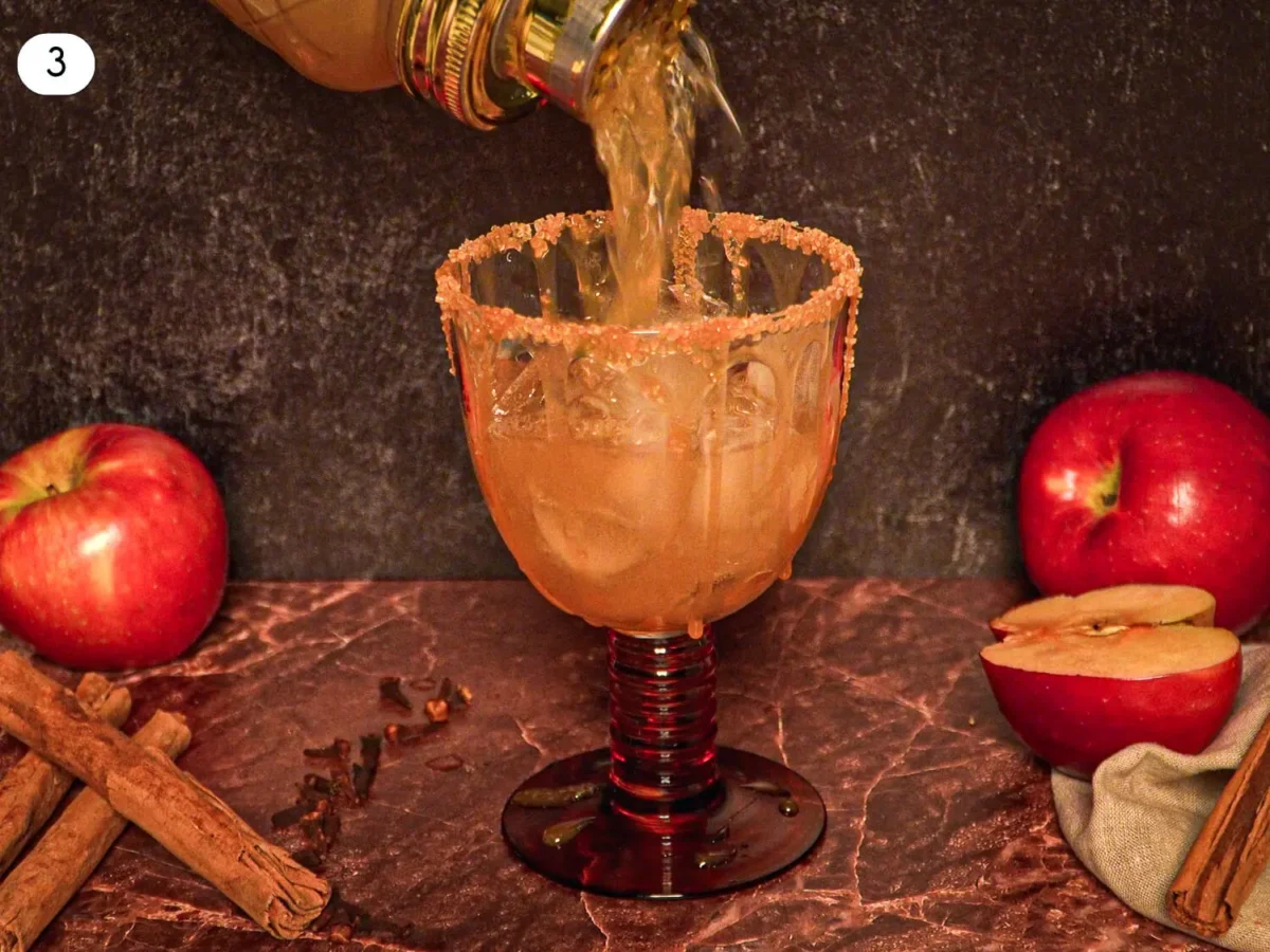 step 3 photo showing an Apple cider margarita mixture being poured over ice into a cinnamon-sugar-rimmed cocktail glass surrounded by apples and cinnamon sticks