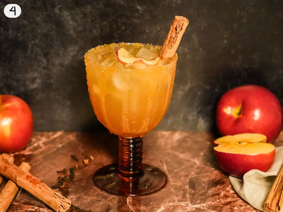 step 4 showing an Apple cider margarita served over ice with a cinnamon-sugar rim, dried apple slice, and cinnamon stick, surrounded by fresh apples