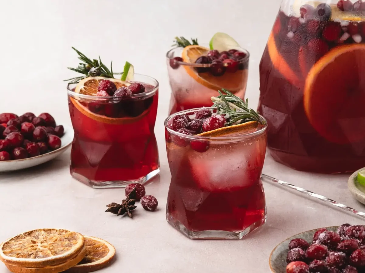 Three cranberry spritzers over ice, garnished with sugared cranberries, orange slices, lime, and rosemary