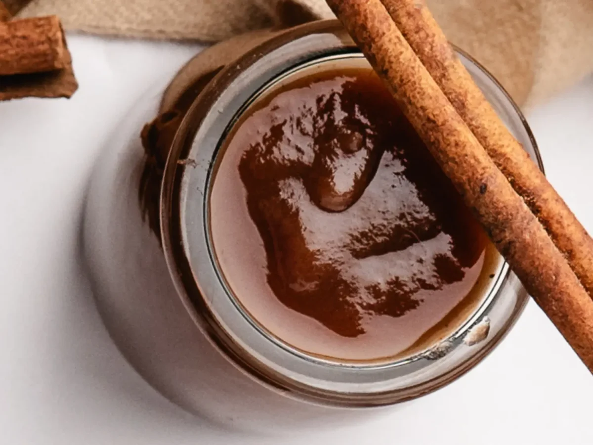 overhead photo of cooked apple butter in a glass jar with a cinnamon stick on top